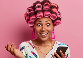 Surprised cheerful ethnic housewife wears hair rollers for making perfect curls dressed in domestic silk robe holds modern cellular poses against pink background. Beauty and hairstyling concept Roller sets